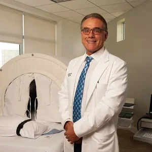doutor marc abreu está de pé, sorrindo. Veste um jaleco branco e gravata azul. Está em um consultório no Instituto Médico BTT